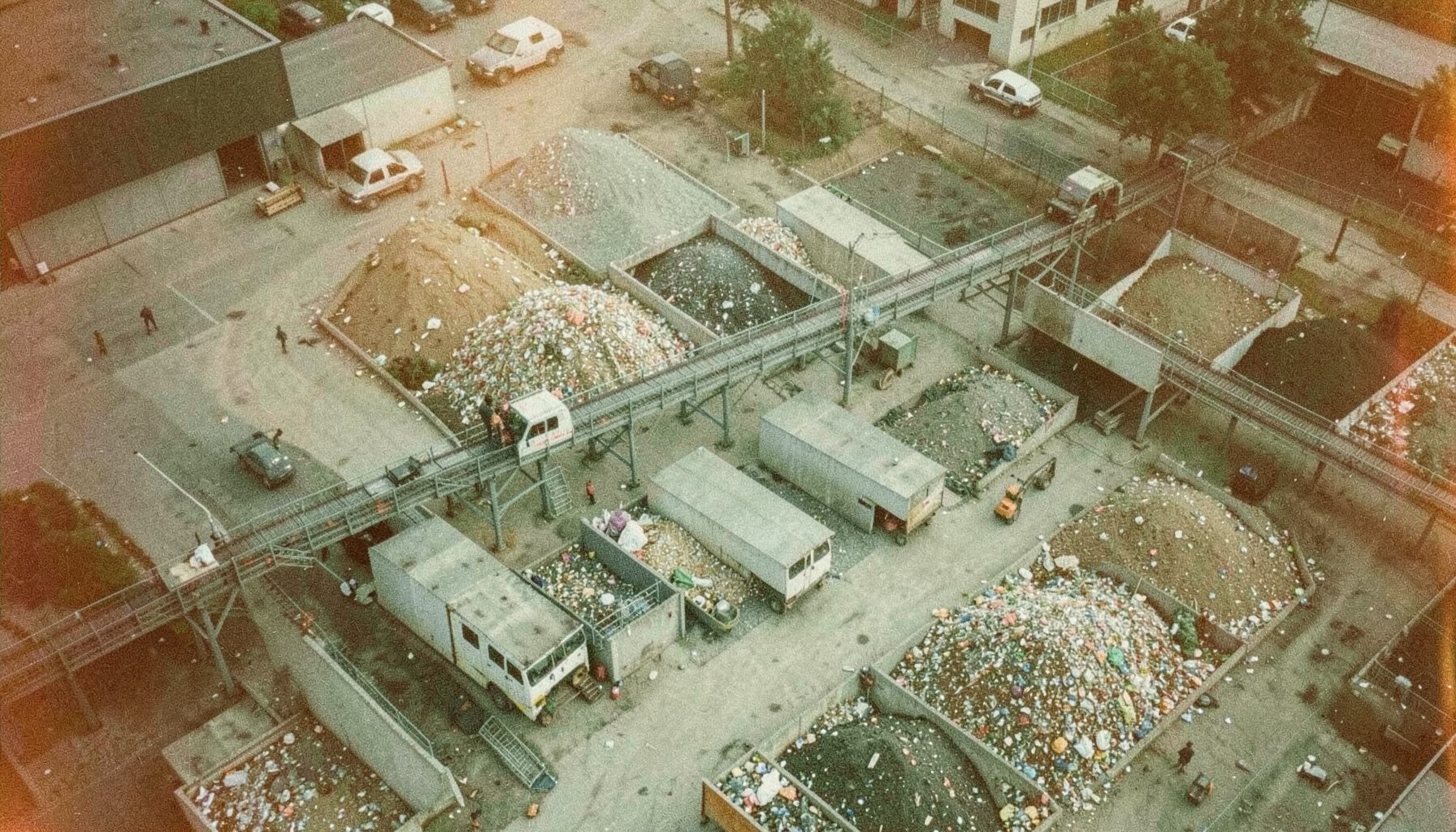 Aerial view of a municipal waste sorting facility with conveyor belts and separated material piles, in muted industrial grays and greens